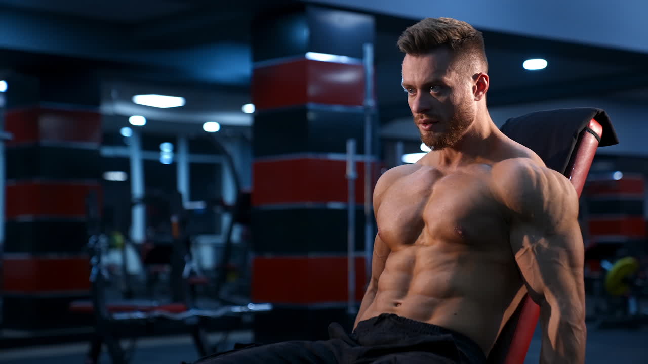 Shirtless young bodybuilder workout with dumbbells. Strong abs. Blue light filter. Video from the side.