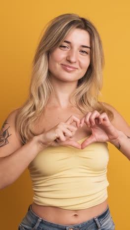 Happy young woman sending flying kiss and showing heart sign with hand on yellow background