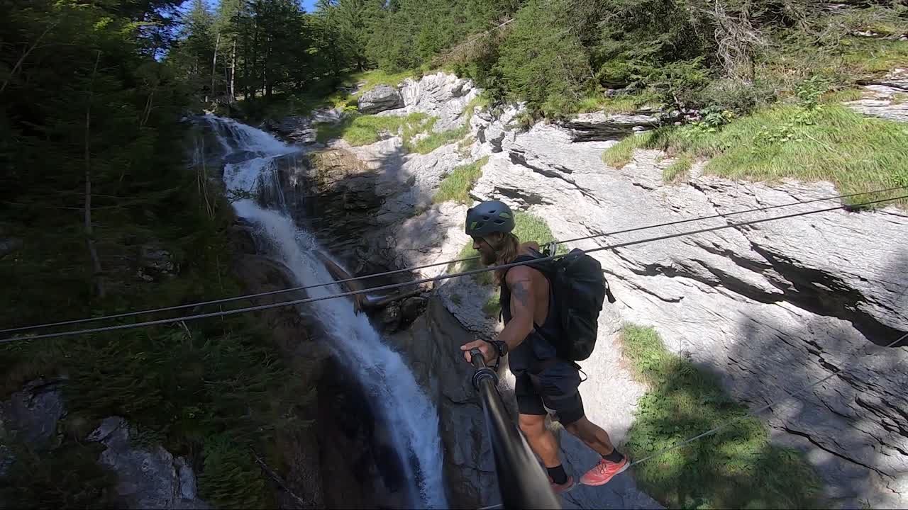 Young man walks on a small and thin line and holding on to another, crossing over a small river and waterfall next to him in the forest of the Swiss alps during a hot summer day. Balancing carefully.