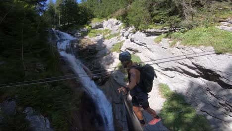 young man walks on a small and thin line and holding on to another, crossing over a small river and waterfall next to him in the forest of the swiss alps during a hot summer day. balancing carefully.