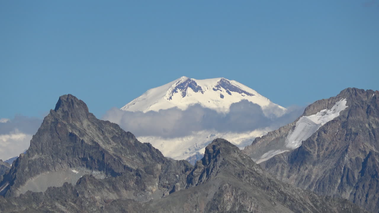 Ampia inquadratura di una vetta innevata che si erge sopra un mare di nuvole e aspre cime rocciose a Dombay, Caucaso, Russia, sotto un cielo azzurro e limpido.
