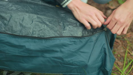 top down view of person adjusting contents of green tent bag while carefully zipping it closed on grassy forest floor, surrounded by pine needles and natural outdoor camping environment