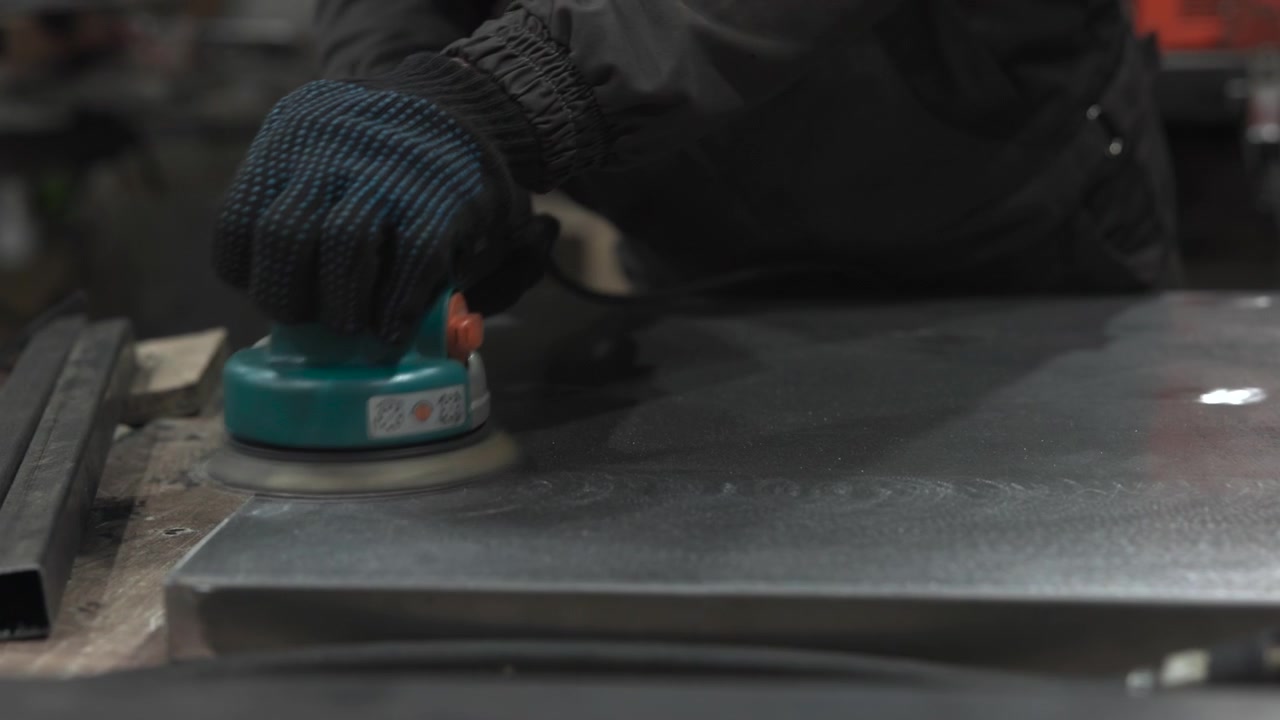 Scena ravvicinata di un'officina industriale mostra mani guantate che guidano una levigatrice orbitale compatta su una lastra di metallo liscia, con corpo dell'utensile color verde acqua, acciaio riflettente, sfondo scuro sfocato e luce fredda e fioca.