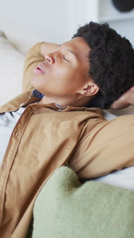 video vertical de un feliz hombre afroamericano relajándose en el sofá con los ojos cerrados en una habitación soleada