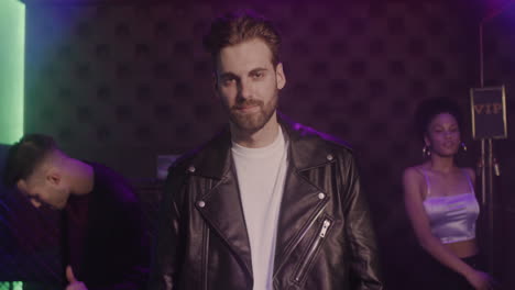 retrato de un apuesto hombre barbudo con chaqueta de cuero sonriendo a la cámara en la discoteca mientras sus amigos bailan detrás de él 1