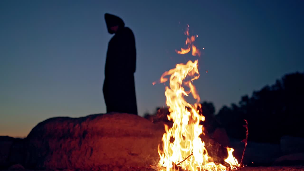 Silhouette of black death on the hill at night. Flame on the ground and dark figure in witch costume in nature. Halloween evening.