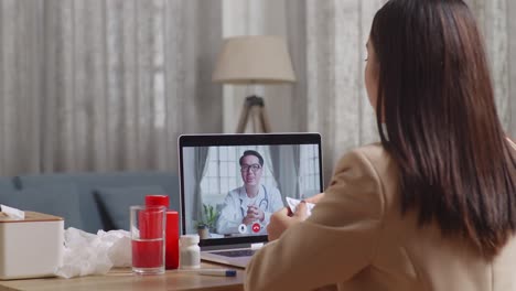 sick asian businesswoman holding a medicine to the video call with doctor on a laptop while working at home