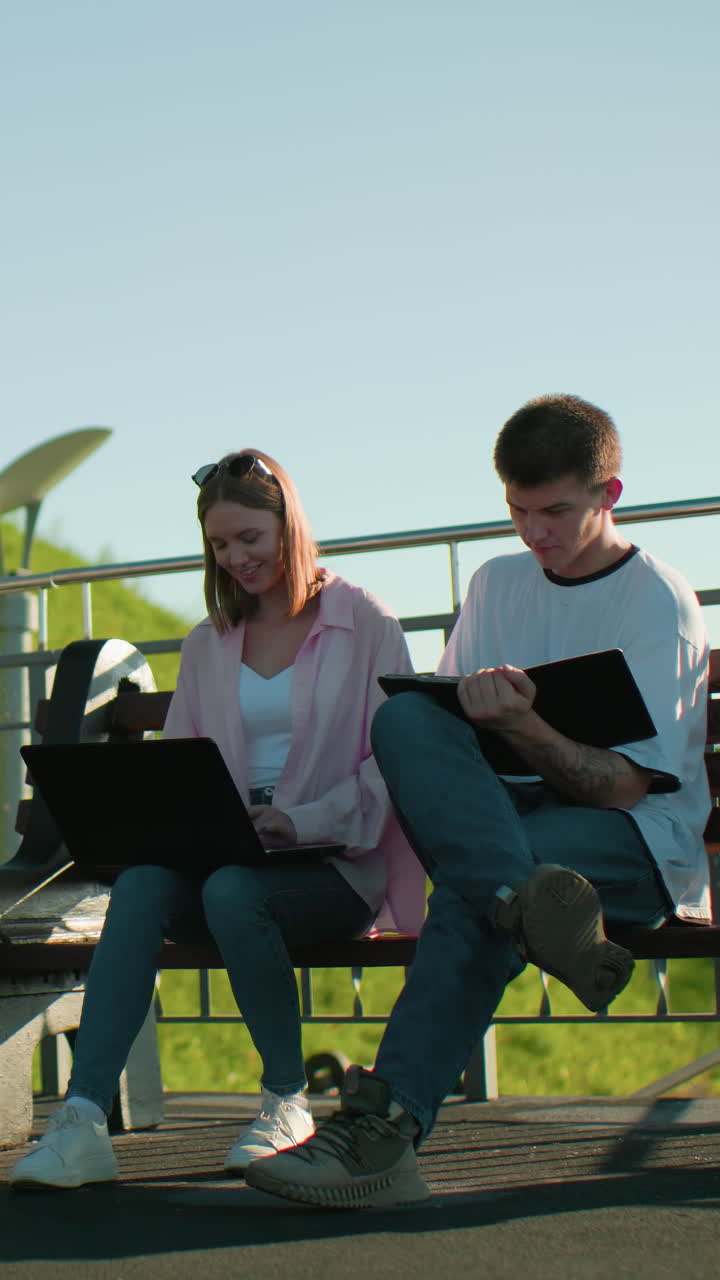 Lady smiling while typing with boy focused on his tablet both seated on a bench, surrounded by greenery and light poles in the background