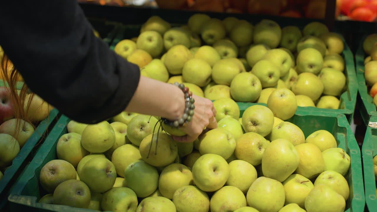 검은 코트와 구슬 팔를 입은 손이 식료품 가게의 상자에서 두 개의 사과를 집어 들고 있는 모습.