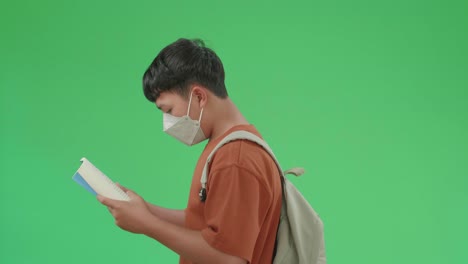 the side view of asian boy student wearing a mask and backpack reading a book while walking to school on green screen chroma key.
