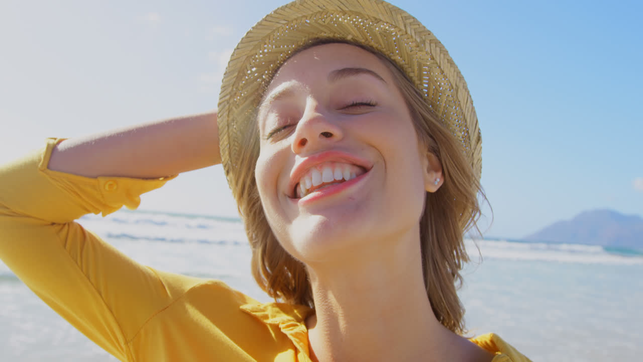 ビーチに立つ帽子をかぶった若い白人女性の正面からの眺め 4K