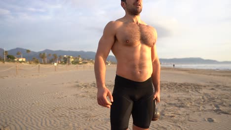 hombre musculoso entrenando con pesas rusas y sin camisa en la playa