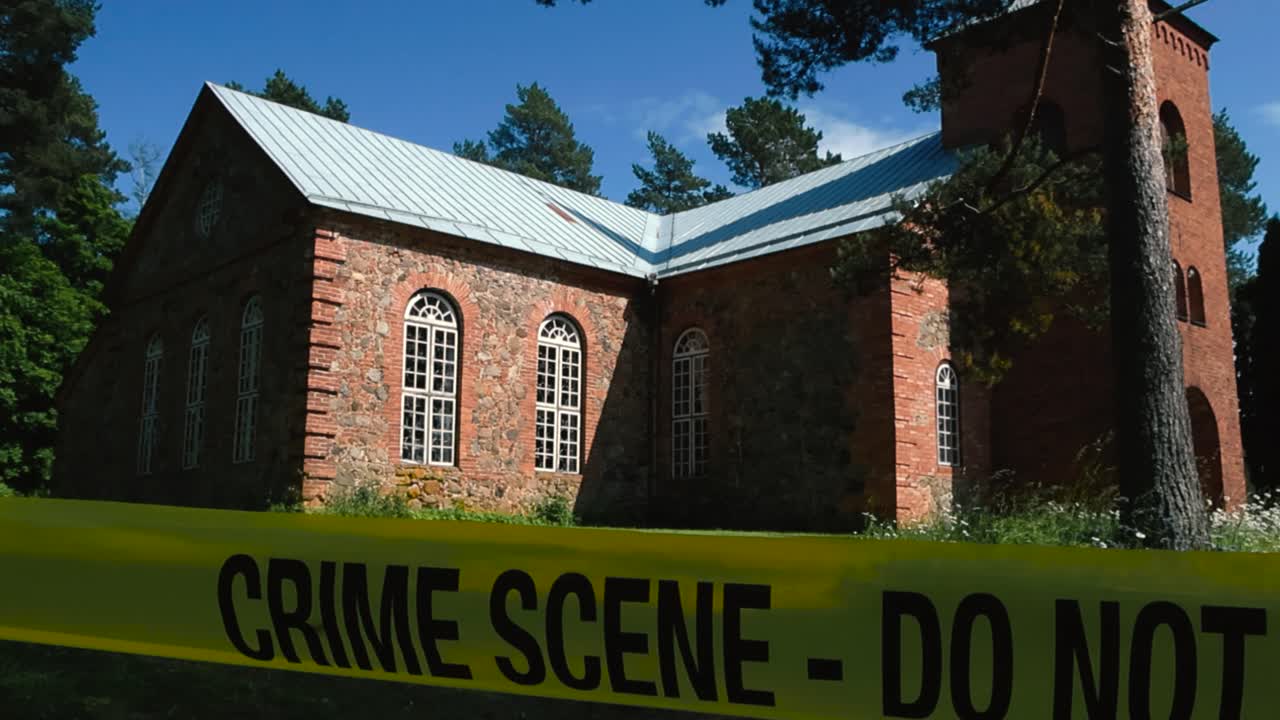 Police Crime Scene tape Do Not Enter in front of a large and old church that is made out of red bricks during a sunny day in a rural area with no people around. Yellow tape is moving in the wind slow.