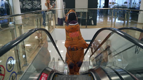 t-rex dinosaur with opened mouth goes up by the escalator. man in inflatable costume of orange dinosaur walking by the shopping mall.