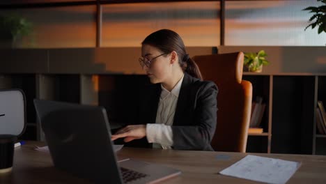 A-concentrated-brunette-girl-in-round-glasses-and-in-a-business-uniform-sits-at-a-wooden-table-and-reviews-sheets-of-papers-with-notes-regarding-the-current-problem-and-its-solution-and-work-plan-in-a-sunny-office