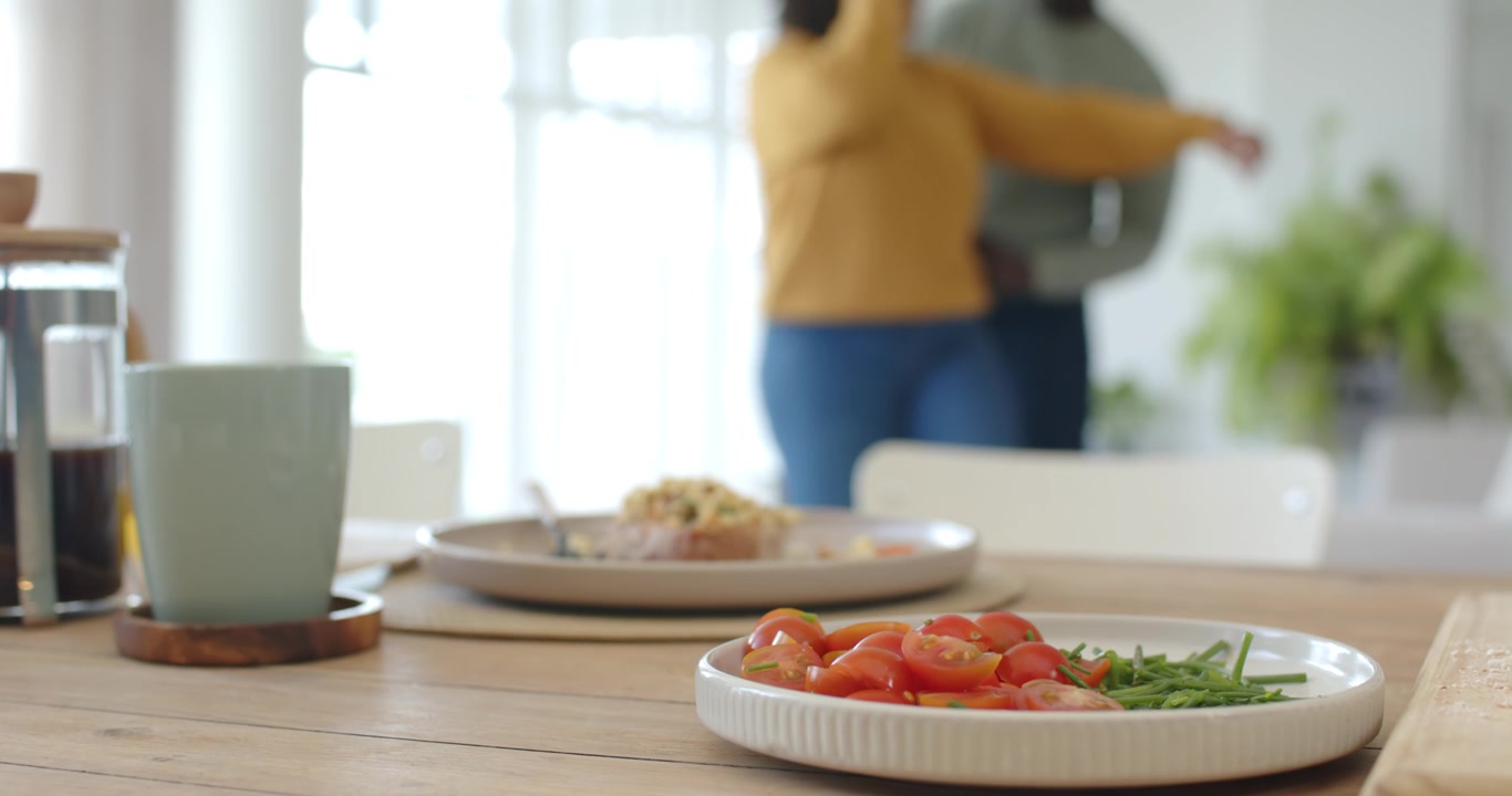 Casal entrando pela direita, abraçando-se e balançando atrás da mesa de jantar, com prato e caneca, permanecendo apaixonados e atentos.