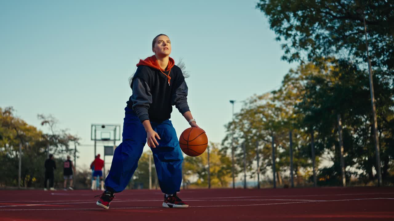 Portrait of a blonde girl in a sports uniform and blue pants who bounces an orange basketball ball off the floor and throws it into the hoop on a red street court in the summer