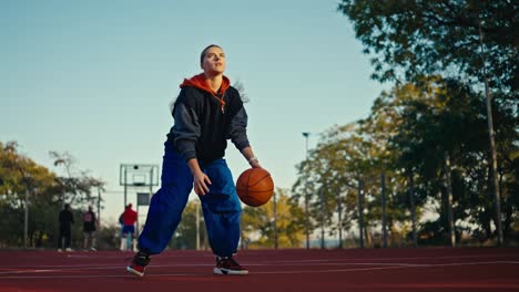 portrait of a blonde girl in a sports uniform and blue pants who bounces an orange basketball ball off the floor and throws it into the hoop on a red street court in the summer