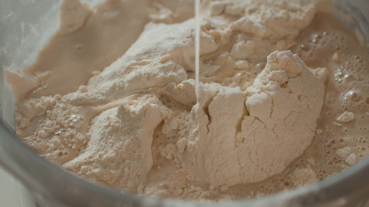 Overhead close-up of flour in stainless steel mixing bowl as thin stream of liquid pours into dry mound, creating darker damp patches, small bubbles, surface cracks, and clumps during dough
