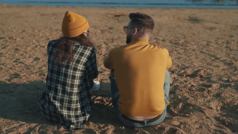 young female and male friends sharing moments sitting on the beach seen from behind