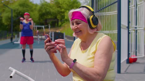 Abuelo-Deportista-Senior-Con-Auriculares-Escuchando-Música-Desde-Un-Teléfono-Móvil-En-El-Patio-De-Juegos