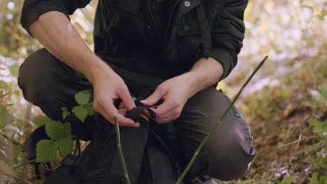 close view of soldier kneeling in forest, carefully placing water bottle in side pouch of bag and securing buckle while small flies land on trousers among surrounding green vegetation and dry leaves