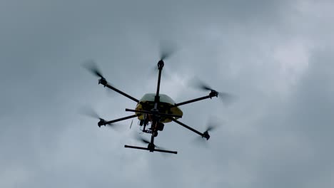 from below, handheld camera captures black and yellow drone with a camera, six propellers, soaring against a cloudy sky