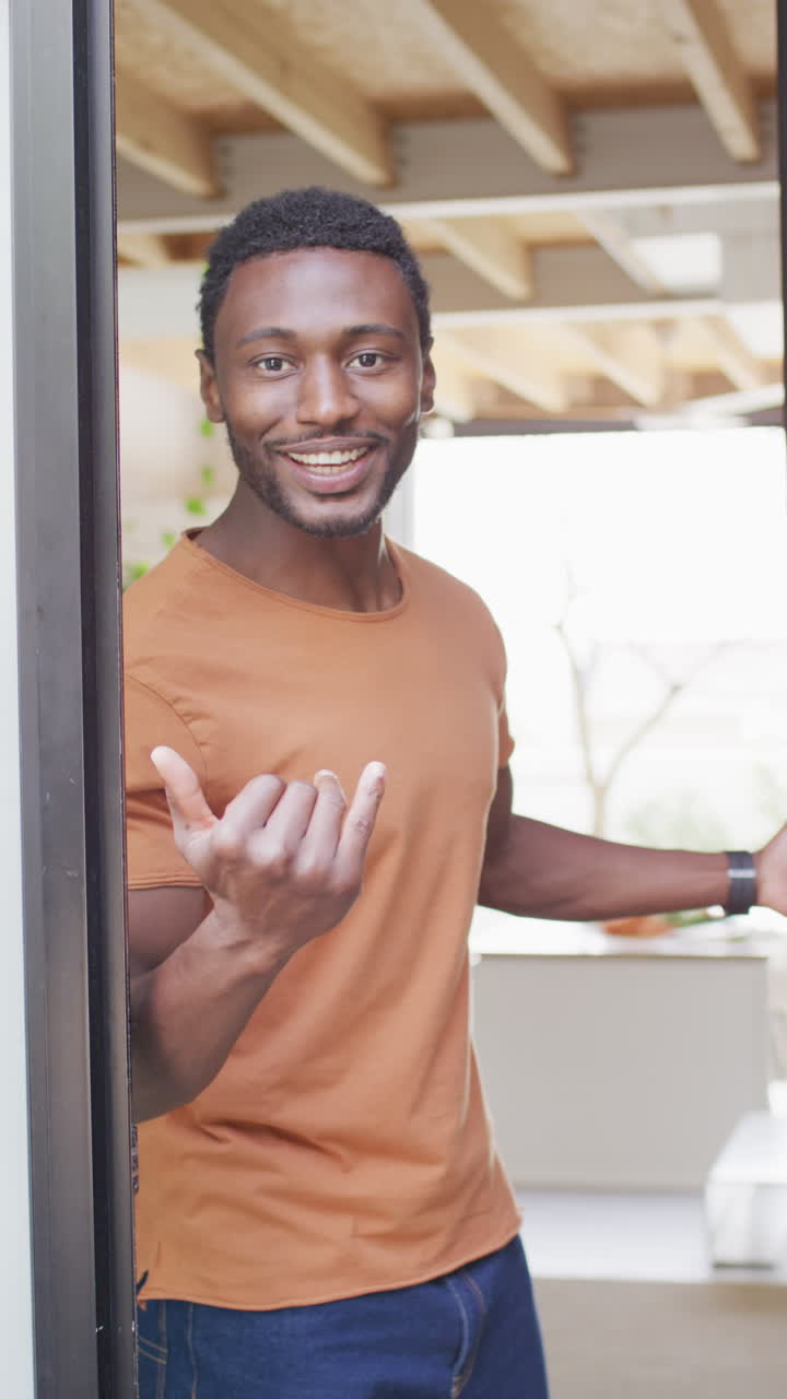 video vertical de un feliz hombre afroamericano de pie en la puerta y invitando dentro de la casa