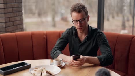 Lindo-Hombre-Rizado-Con-Anteojos-Sentado-En-Un-Café-Toma-Un-Teléfono-Inteligente