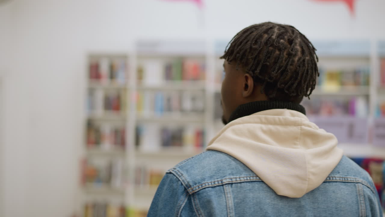 Back shot of man with dreadlocks dressed in denim jacket and hoodie, browsing books in store, with colorful bookshelves and bright lighting in background, calm and relaxed atmosphere