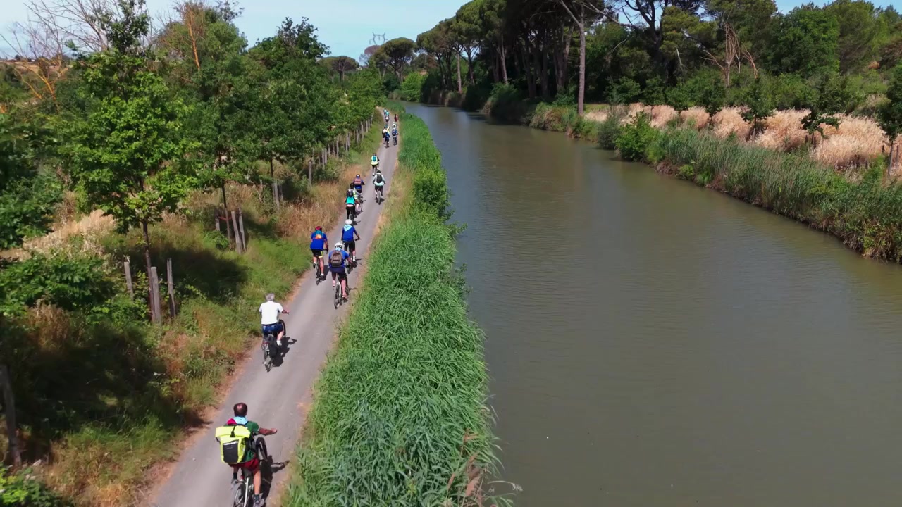 Gruppe af cyklister cykler langs Canal du Midi i frodig landlig vegetation, dronefølger over rytterne.