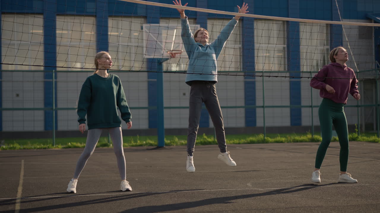 후디를 입은 여성들이 야외에서 배구 훈련에 참여하고, 코트에서 연습하고, 배경은 건물과 스포츠 시설을 보여줍니다.