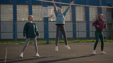 mujeres con capuchones participan en una sesión de entrenamiento de voleibol al aire libre, practicando en la cancha, el fondo muestra el edificio y la instalación deportiva, capturado en clima soleado