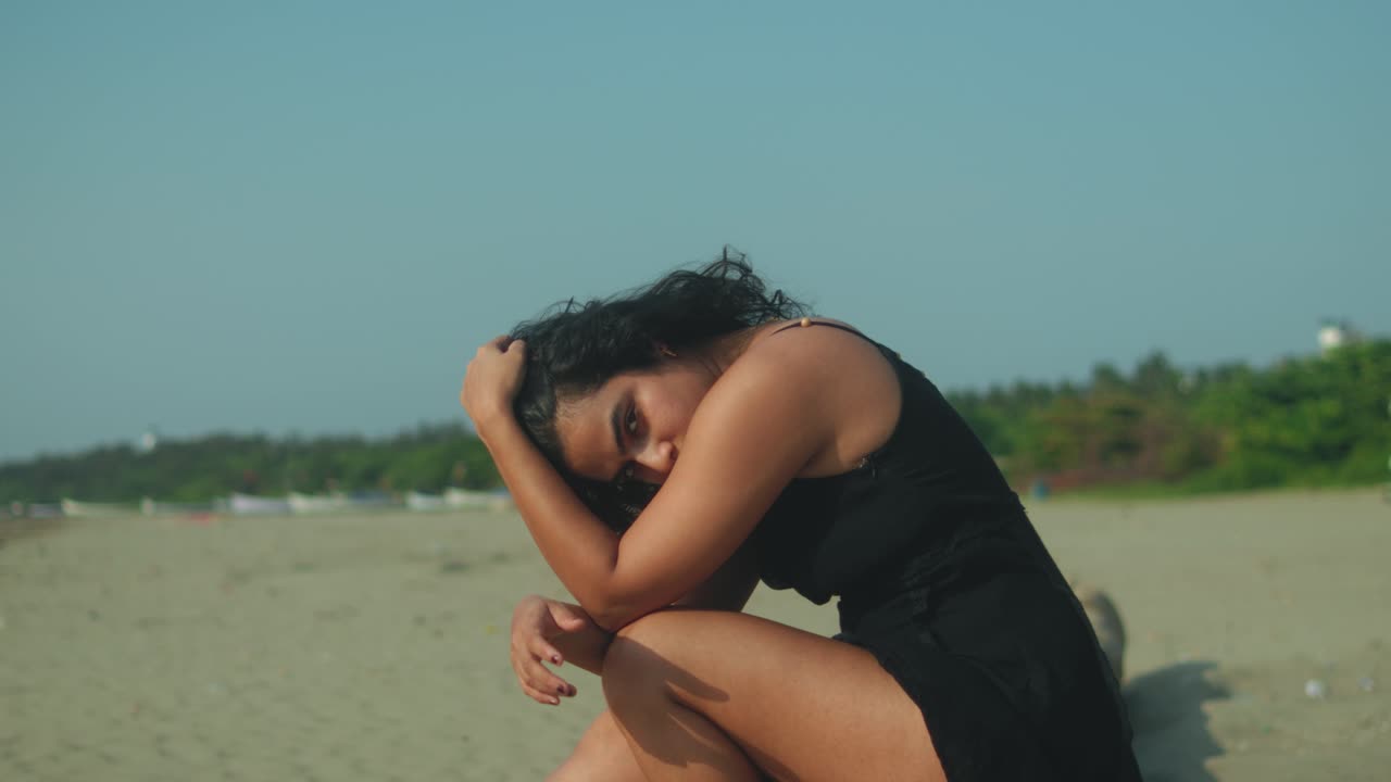 A woman in a black dress sits on the sand, resting her hand on her head, lost in thought on a deserted beach under clear sky. Scene conveys solitude, calm, and reflection by the sea, slow motion shot