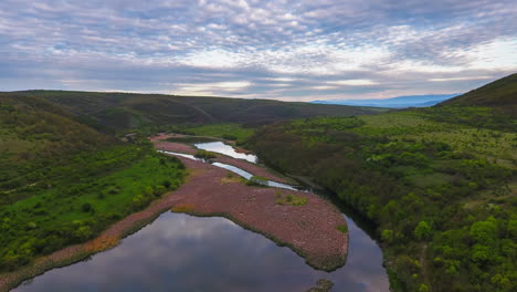 Vista-Aérea-En-Time-lapse-De-Un-Pintoresco-Valle-Fluvial-Al-Anochecer-Con-Colinas-Onduladas-Y-Un-Cielo-Nublado-Colorido