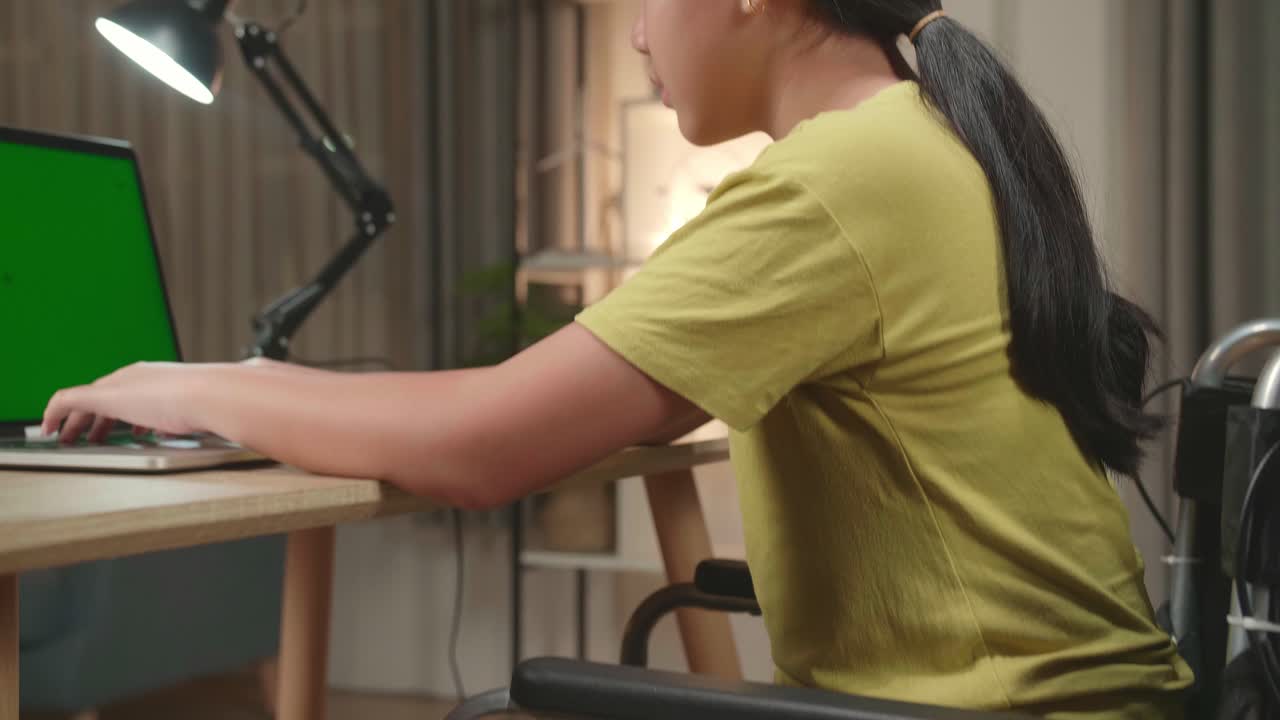Asian Kid Girl Sitting In A Wheelchair Using Laptop Computer With Mock Up Green Screen And Pain Her Back At Home
