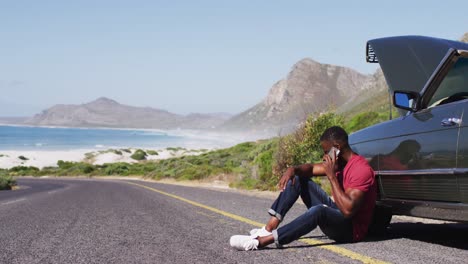 african american man talking on smartphone while standing near his broken down car on road