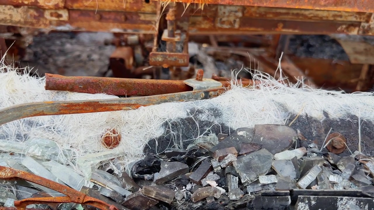 Primo piano macro di detriti carbonizzati, filamenti di fibra di vetro fusi e parti metalliche arrugginite all'interno dell'abitacolo di un autobus gravemente danneggiato da un incendio!
