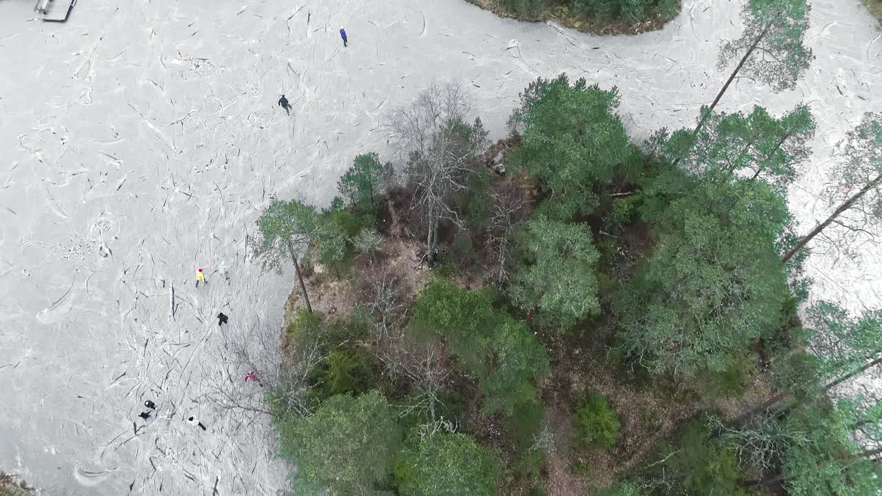 Aerial view showing group of happy people ice skating on frozen lake around trees growing on island - Tilt up movement