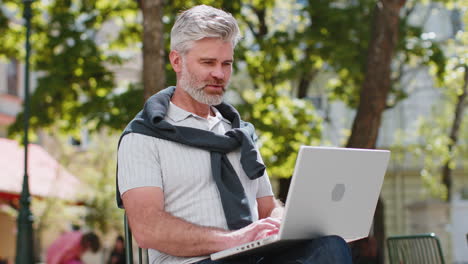 hombre de mediana edad trabajando en una computadora portátil envía mensajes hace compras en línea sentado en la calle de la ciudad