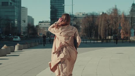 la cámara sigue a una hermosa mujer con un elegante abrigo de otoño beige claro caminando sola por la gran plaza soleada de la ciudad en cámara lenta.