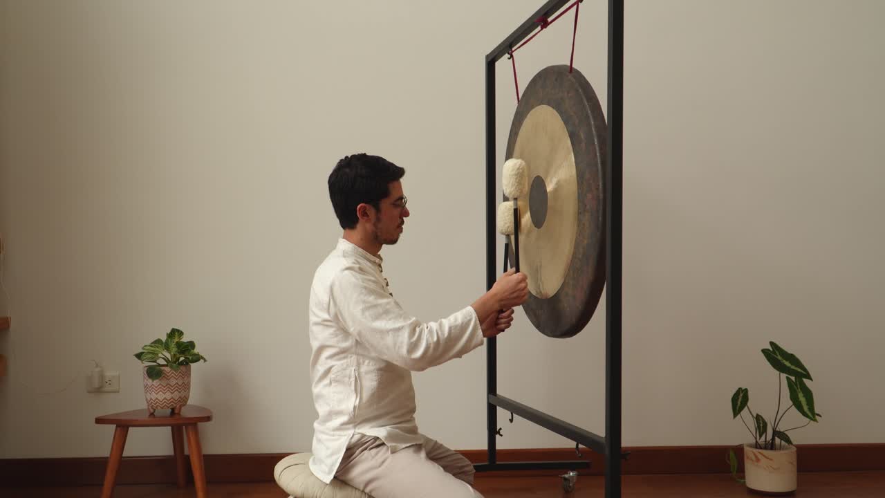 Side-view of a man kneeling on a cushion and playing a large gong with two soft mallets in a calm minimalist studio with plants—ideal for wellness, meditation, sound healing and therapy themes