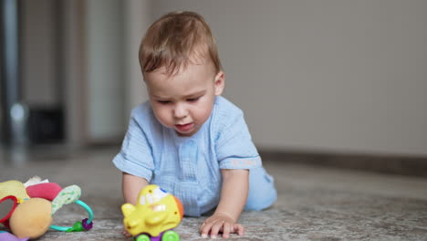 nice cute baby boy in blue clothes stands on all fours on the floor. lovely kid tries to choose a toy in front of him. blurred backdrop.
