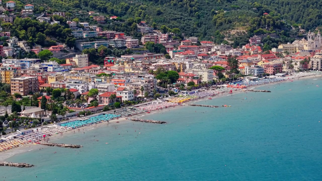 Ripresa aerea dall'alto con drone che si allontana dal Mar Mediterraneo verso Laigueglia, rivelando il lungo litorale, la città balneare, le barriere frangiflutti e le verdi colline liguri