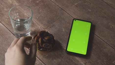 Toma-En-ángulo-Alto-De-Agua-Y-Dátiles-En-Una-Mesa-De-Madera-Con-Teléfono-De-Pantalla-Verde