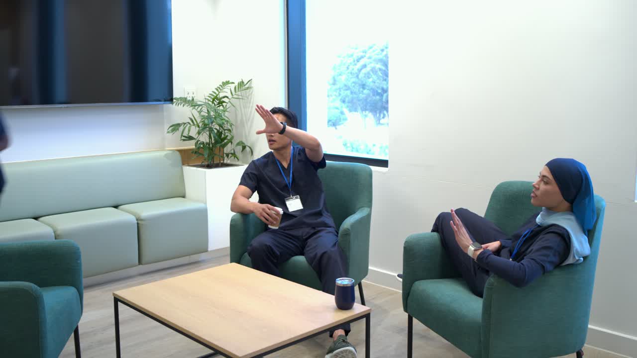 Equipo diverso hablando en una sala de descanso, un hombre asiático con una taza gesticulando, un compañero que le entrega un teléfono