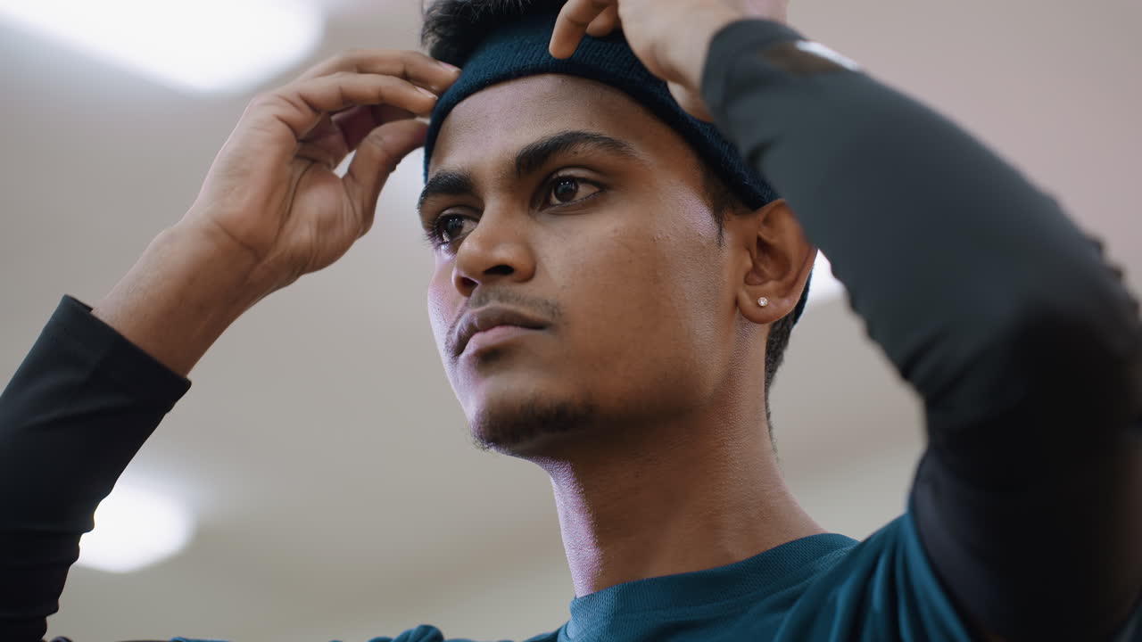 Close up of athlete with earring adjusting blue headband under fluorescent indoor light, focused expression, blurred background, preparation before training