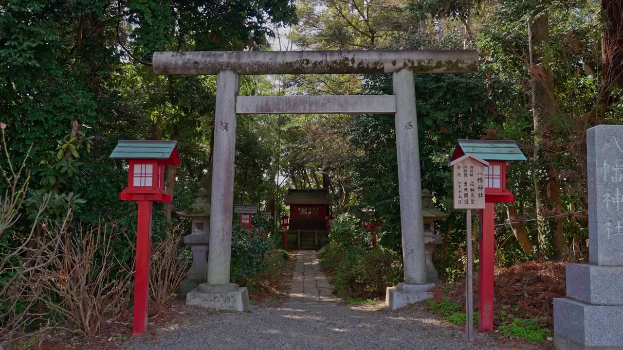 A weathered stone torii gate flanked by traditional red lanterns stands at the entrance of a tranquil Shinto shrine, framed by dense green trees and leading to a vermilion sanctuary beyond.