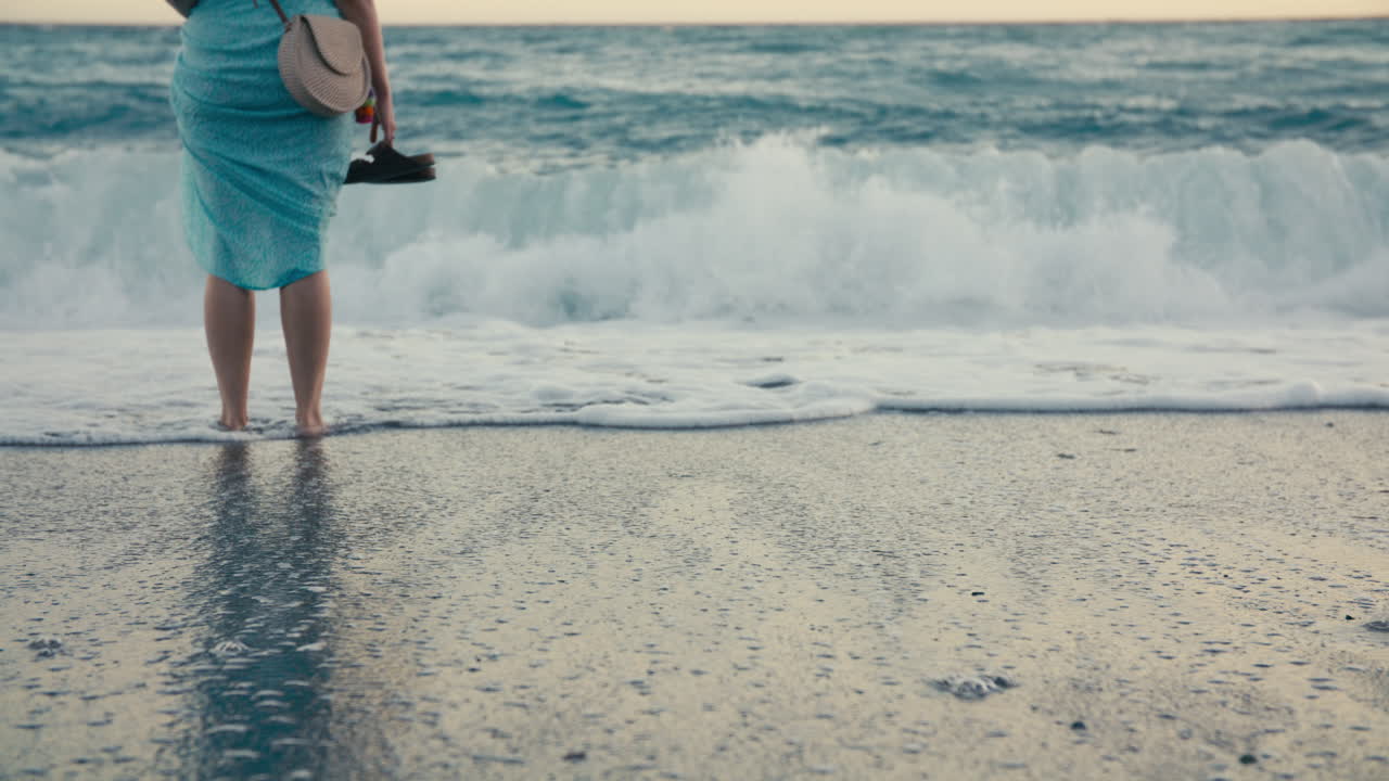 A young woman stands barefoot on the beach, holding her sandals as waves crash ahead and covers her feet. Golden light reflects in the wet sand