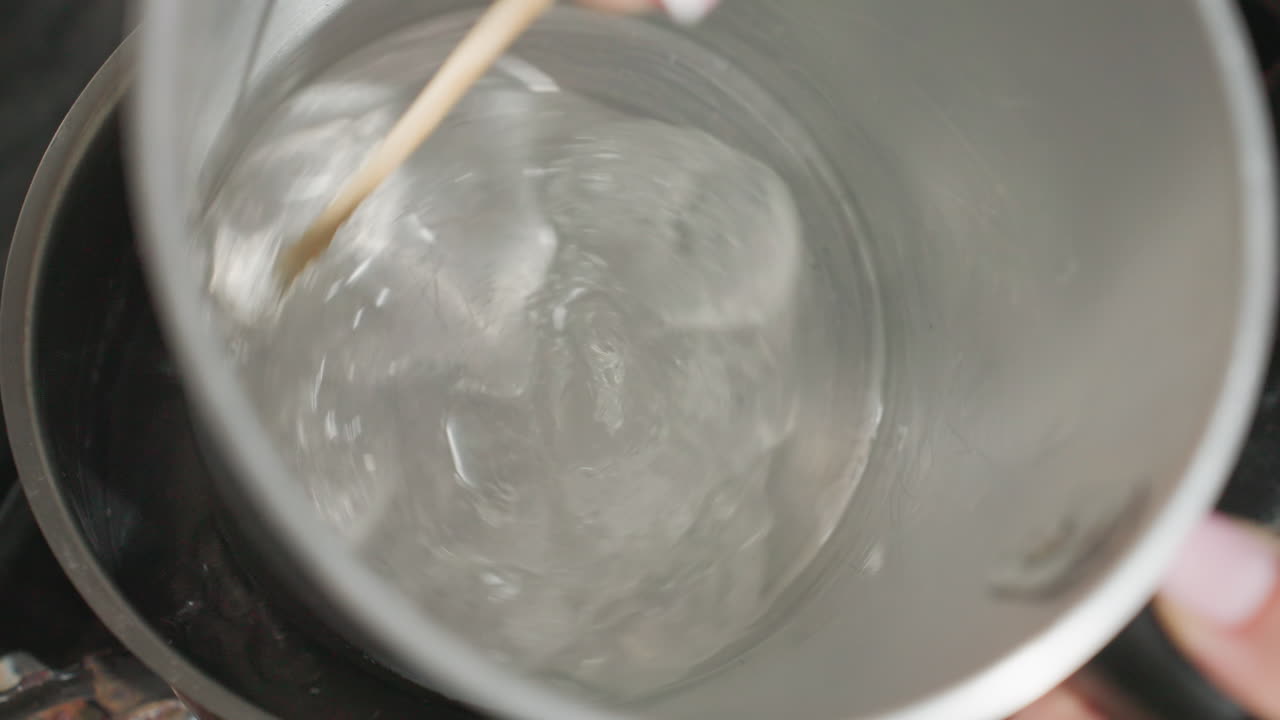 Close up of person stirring soluble mixture inside metal jug placed in steaming pot on gas burner, creating swirling motion in hot water as heat rises during kitchen preparation process